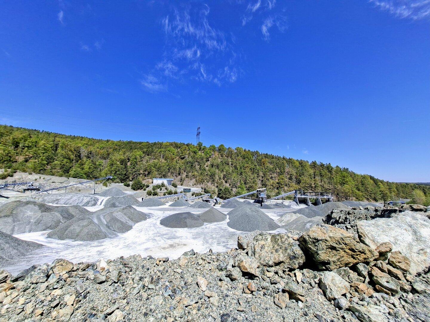 Steinbruch mit mehreren Kieshügeln, Förderbändern und angrenzendem Wald unter klarem, blauem Himmel.
