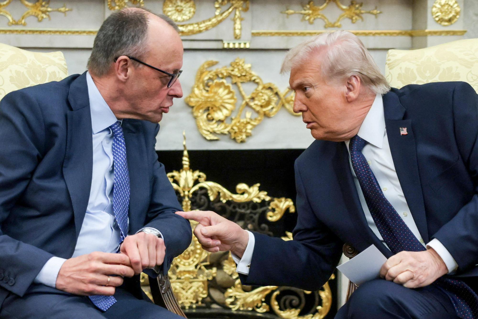 Zwei Männer in Anzügen unterhalten sich vor einem goldenen, verzierten Hintergrund.