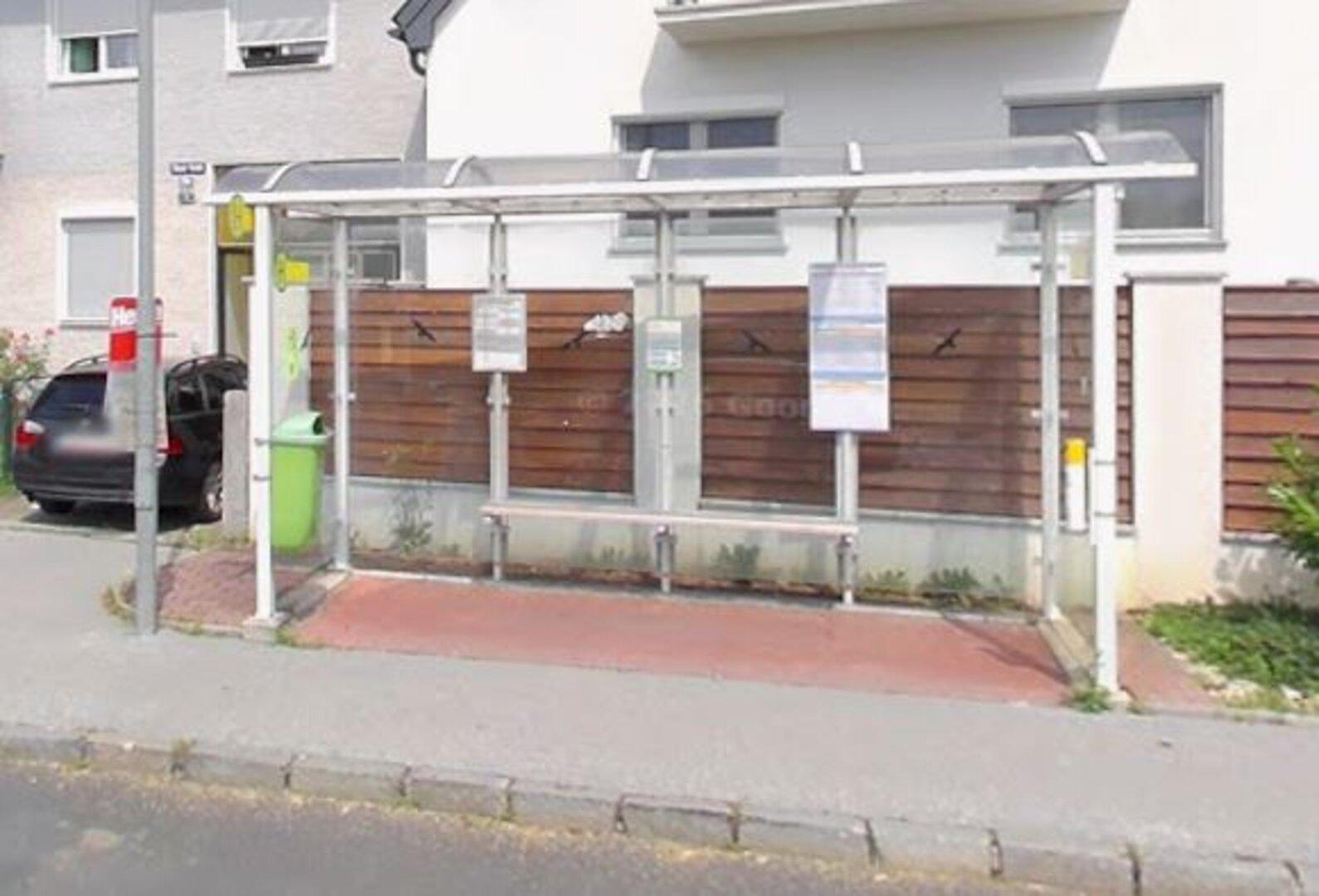 Überdachte Bushaltestelle mit Sitzbank, Fahrplänen, grünem Mülleimer und Holzwand im Hintergrund.