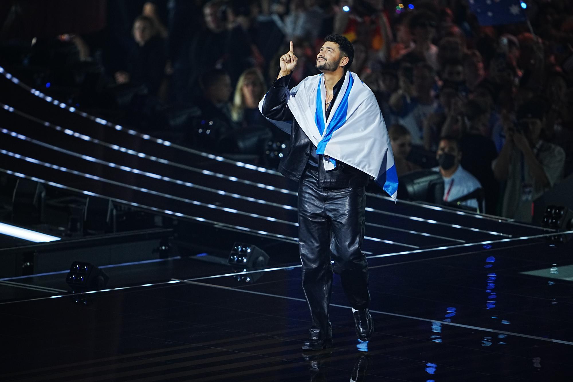 Ein Mann mit einer blau-weißen Flagge um die Schultern steht auf einer Bühne und zeigt mit dem Finger nach oben.