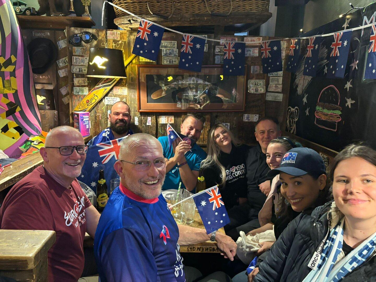 Eine fröhliche Gruppe von Menschen sitzt in einem dekorierten Raum mit vielen australischen Flaggen und lächelt in die Kamera.