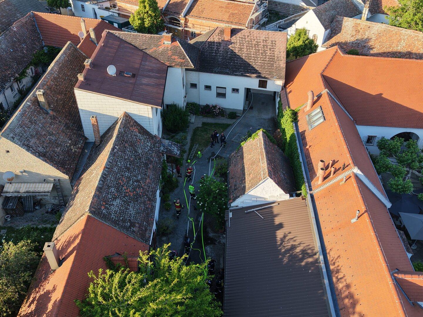 Mehrere Feuerwehrleute stehen in einem Innenhof zwischen Häusern mit roten Dächern und arbeiten mit Schläuchen und Leitern.