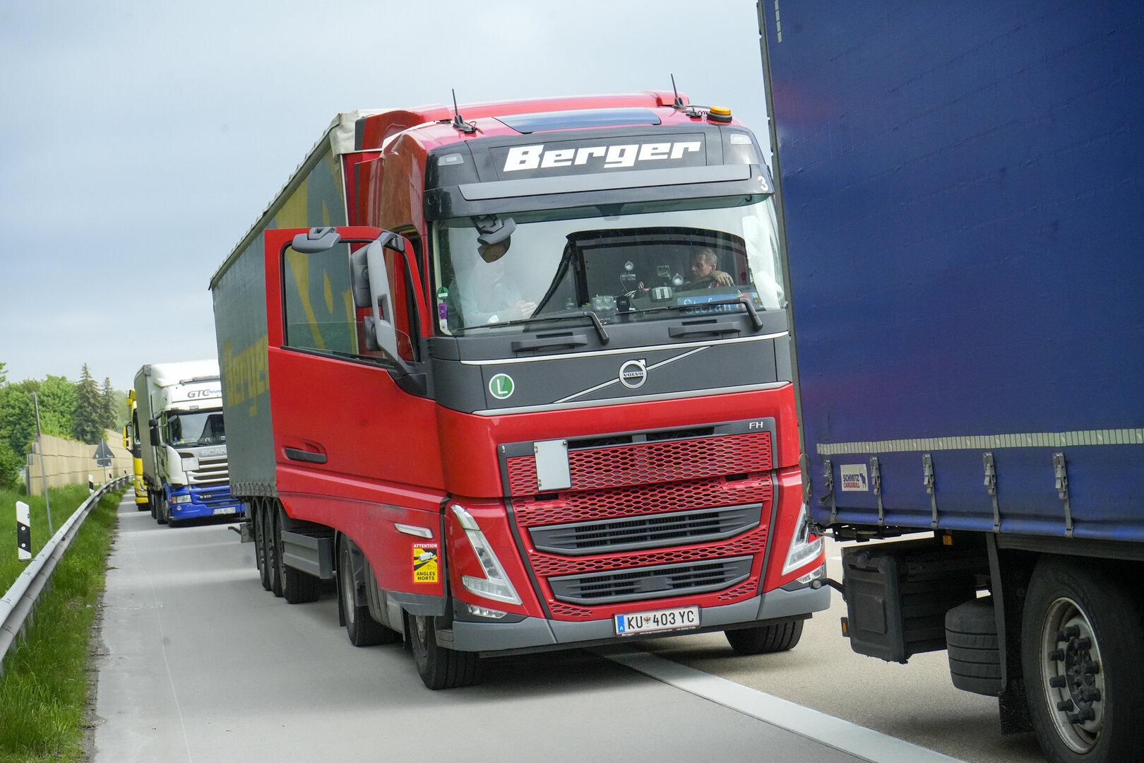 Ein roter Lkw fährt dicht neben einem blauen Lkw auf einer Autobahn, weitere Lkw sind im Hintergrund zu sehen.