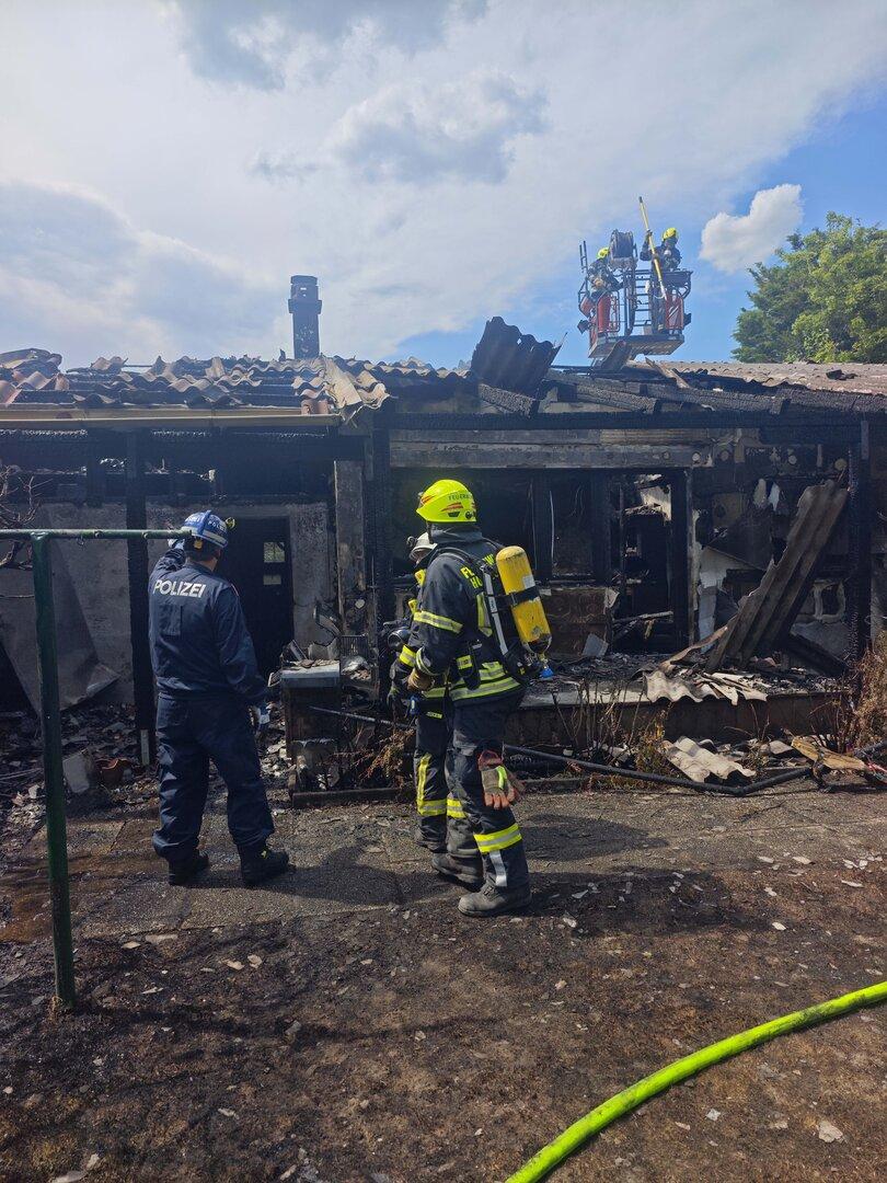 Zwei Feuerwehrleute und eine Polizistin stehen vor einem ausgebrannten Gebäude, während weitere Einsatzkräfte auf einer Drehleiter arbeiten.