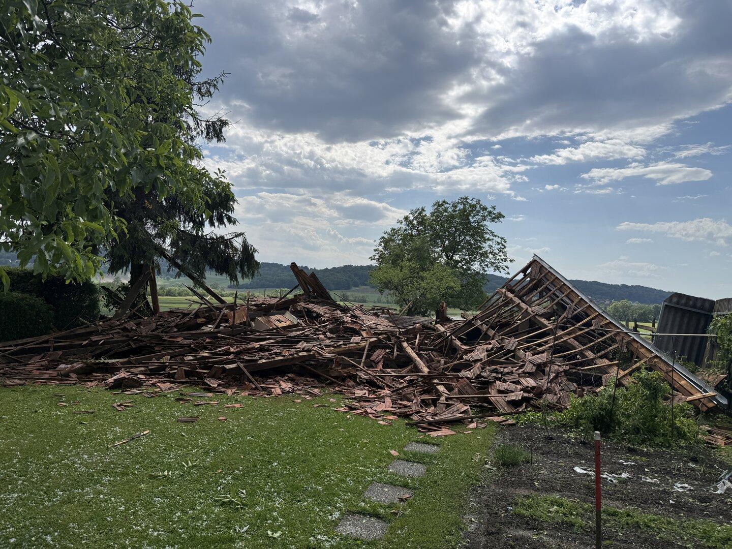 Ein eingestürztes Holzgebäude liegt in Trümmern auf einer Wiese, umgeben von Bäumen und ländlicher Landschaft.