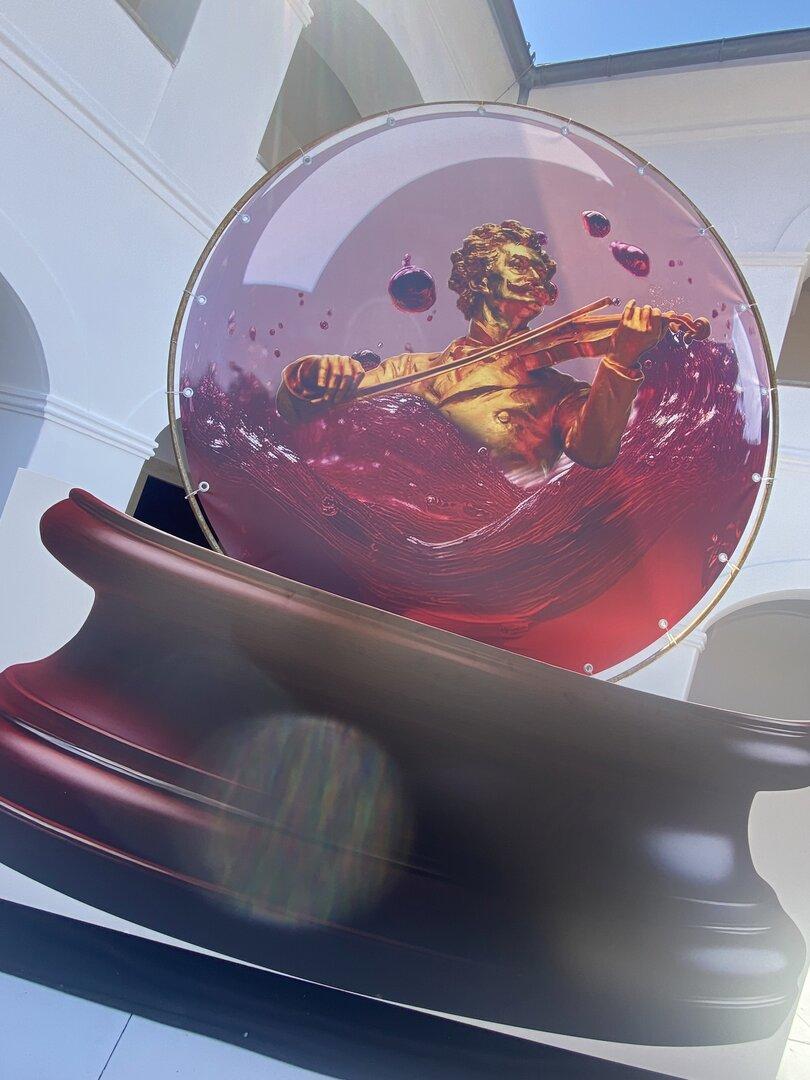 Eine goldene Figur spielt Geige in einer Glaskugel, die mit roter Flüssigkeit und Blasen gefüllt ist.