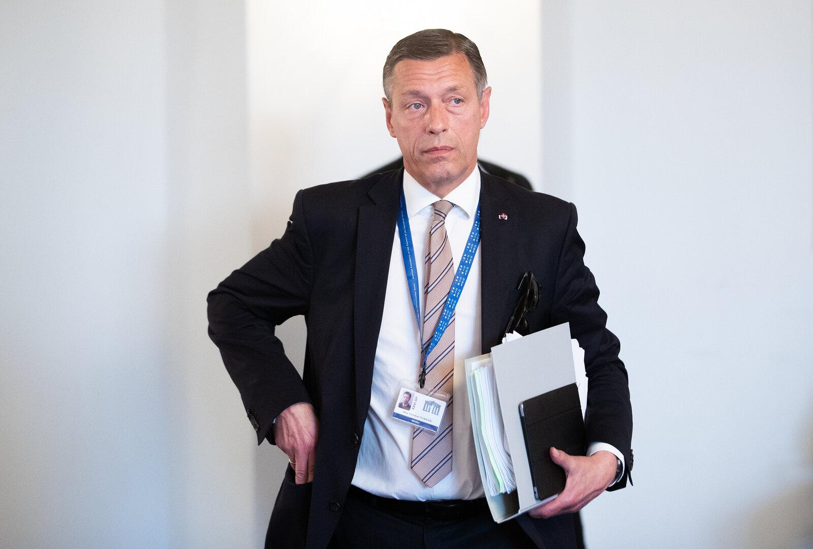 Ein Mann im Anzug mit Akten und Ausweis um den Hals steht vor hellem Hintergrund.