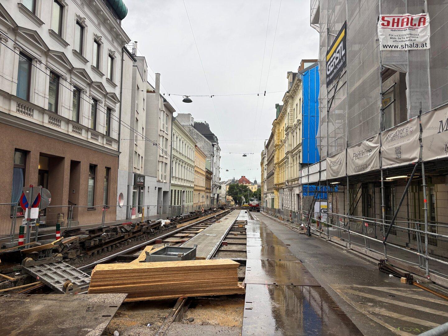 Straße mit Baustelle und offenen Straßenbahnschienen, umgeben von mehrstöckigen Häusern und Baugerüsten bei nassem Wetter.