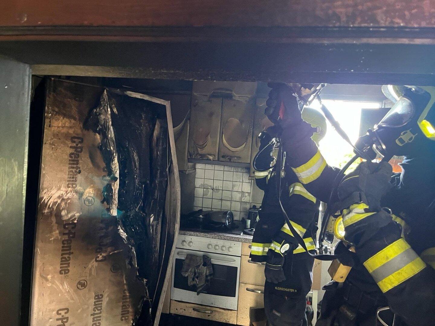 Zwei Feuerwehrleute in Schutzkleidung löschen einen Brand in einer stark beschädigten Küche mit verbrannten Geräten und Möbeln.