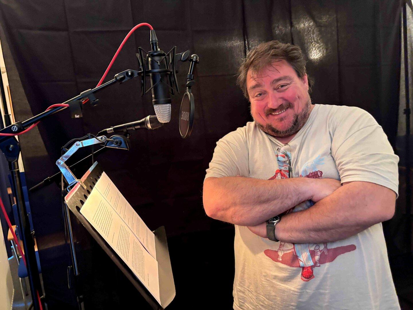 Ein lächelnder Mann steht vor Mikrofonen und einem Notenständer in einem Tonstudio.