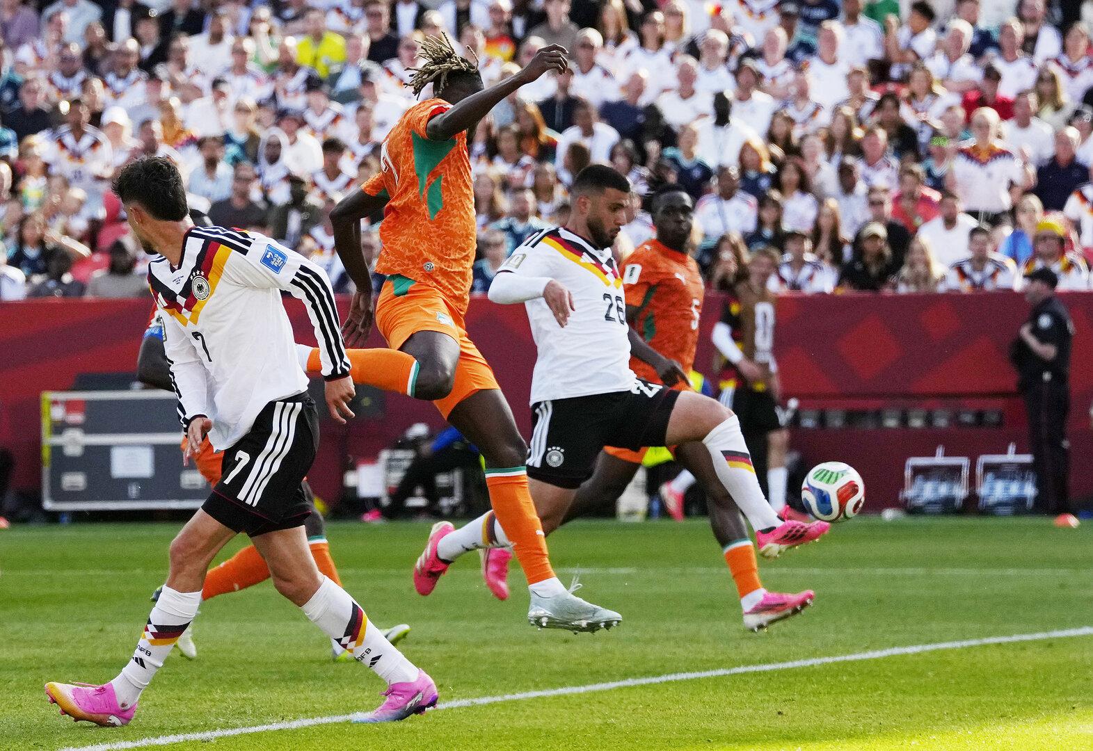 Fußballspiel mit Deutschland gegen die Elfenbeinküste, mehrere Spieler kämpfen um den Ball vor voller Tribüne.