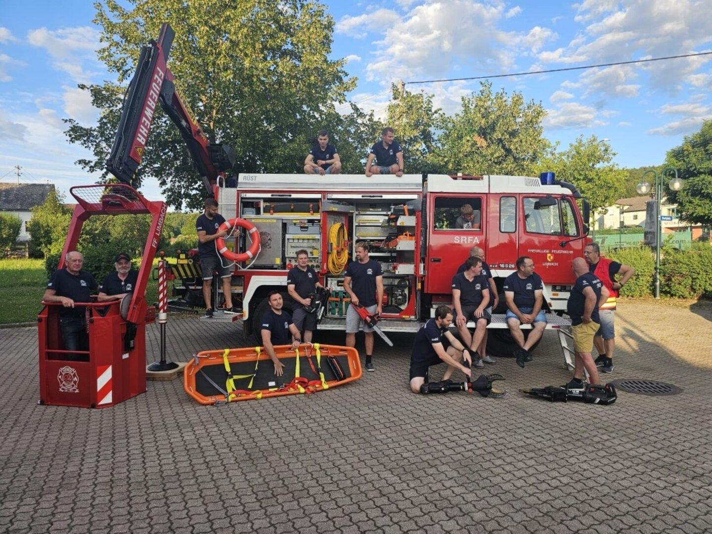 Mehrere Feuerwehrleute stehen und sitzen an einem roten Einsatzfahrzeug mit Rettungsgerät und geöffneter Geräteraumklappe.