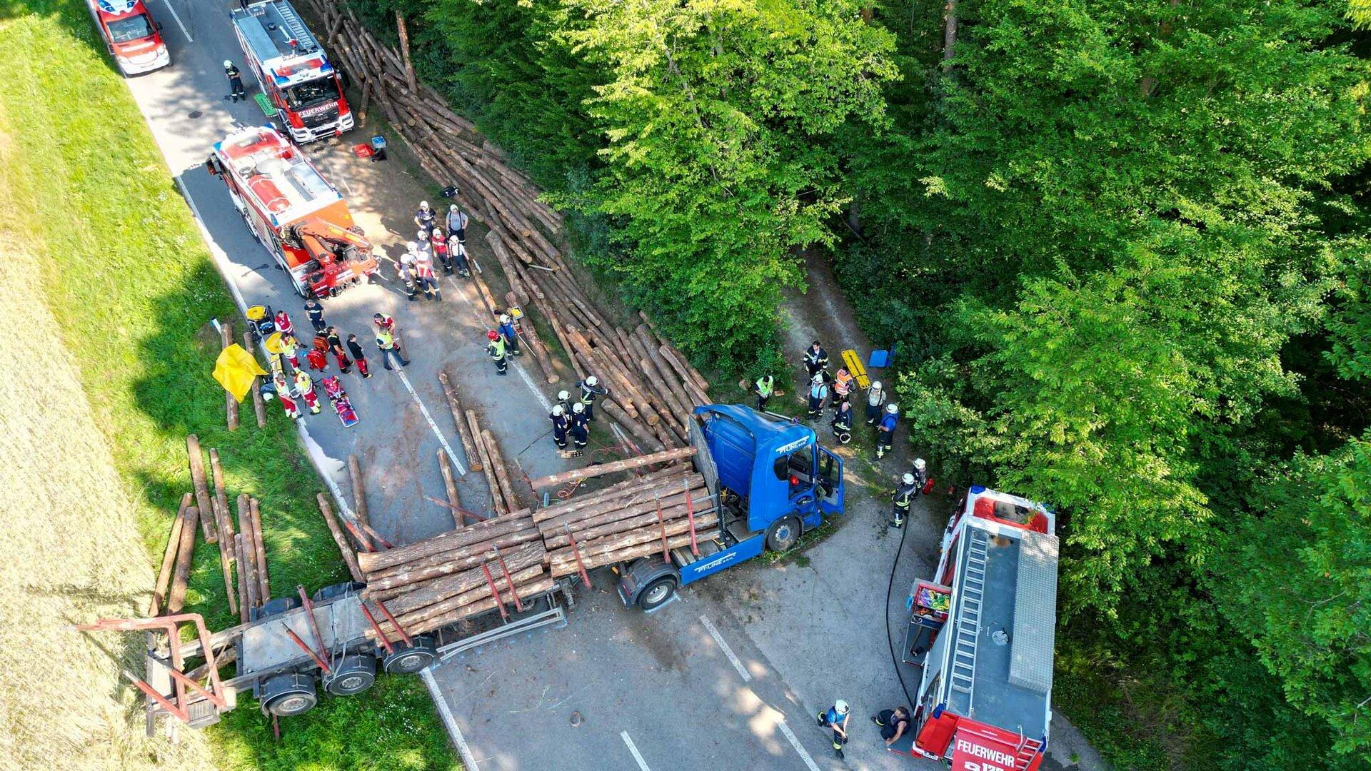 Luftaufnahme eines Unfalls mit umgestürztem Holztransporter, Feuerwehrleuten und mehreren Einsatzfahrzeugen auf einer Waldstraße.
