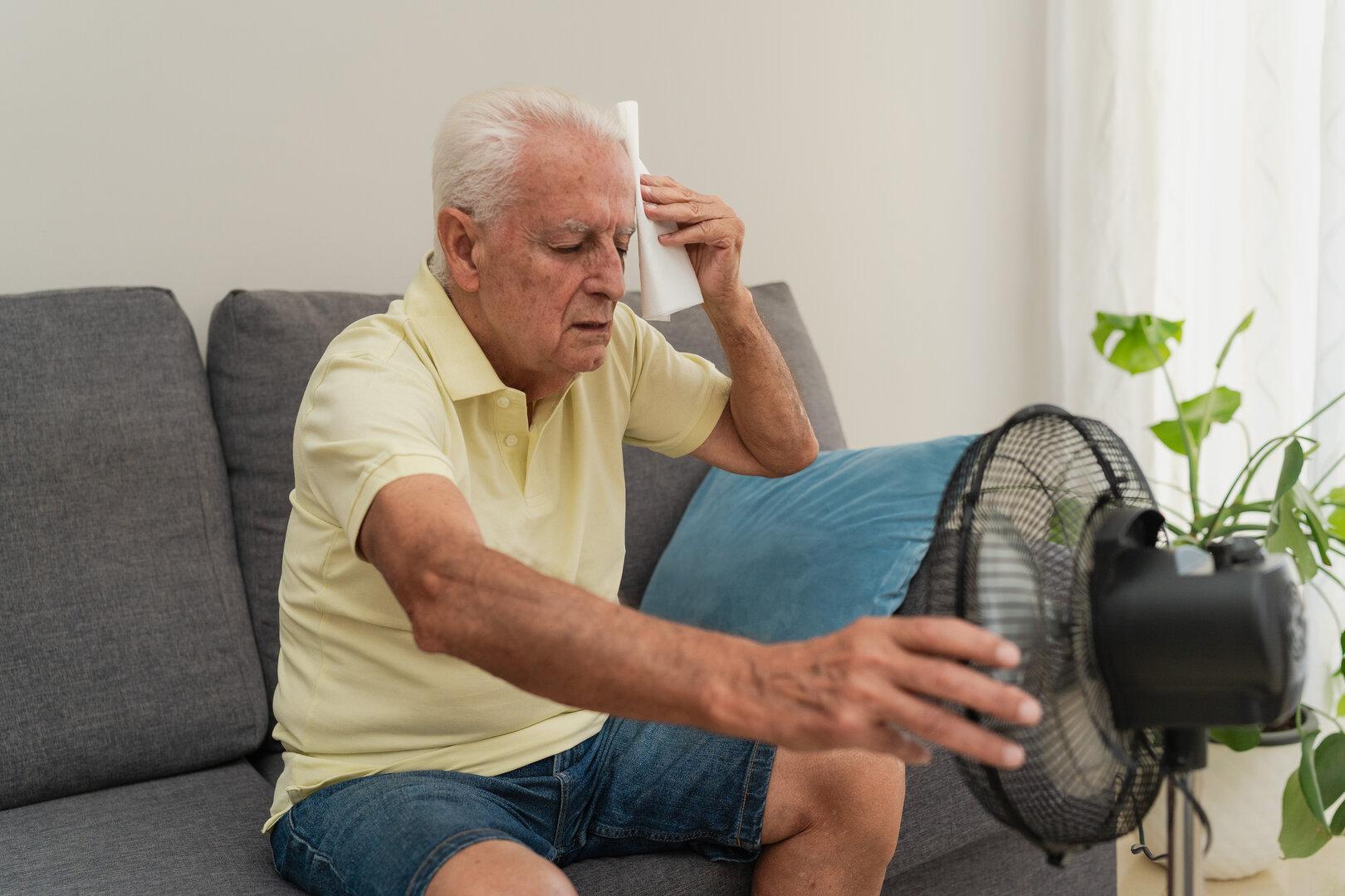 Senior man suffering from summer heat wiping sweat with napkin and holding a electric fan at home
