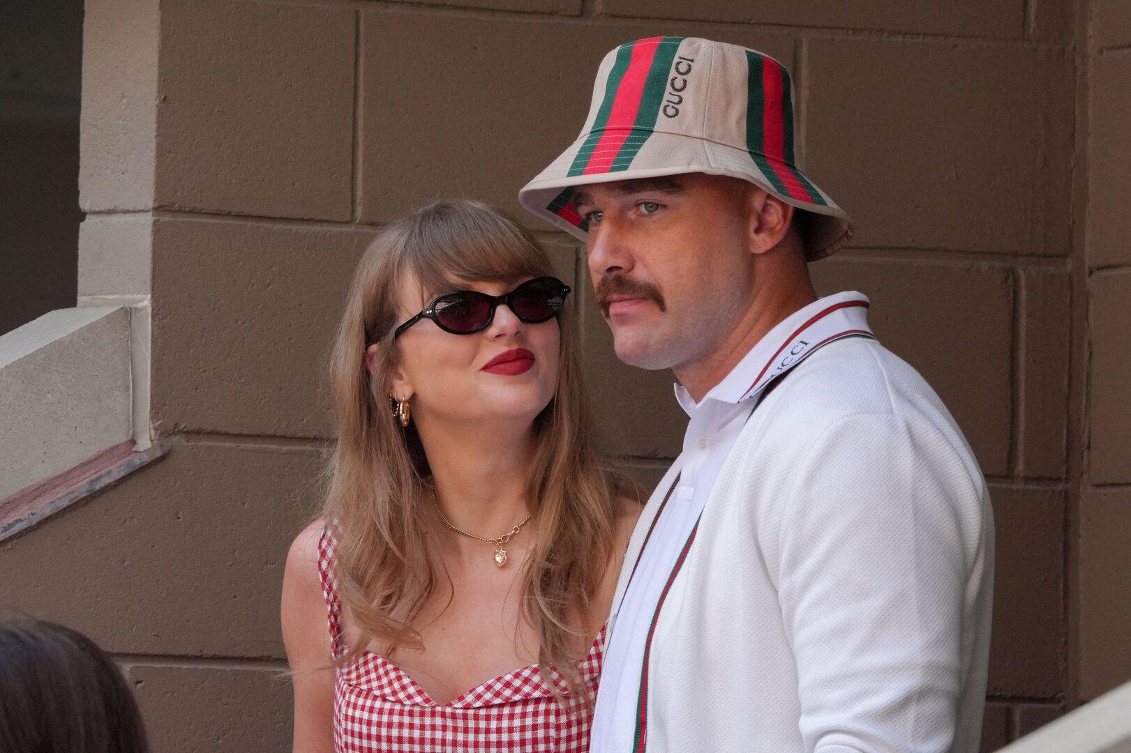 Taylor Swift mit Sonnenbrille blickt zu Travis Kelce, der einen Gucci-Fischerhut trägt, vor einer braunen Ziegelwand.