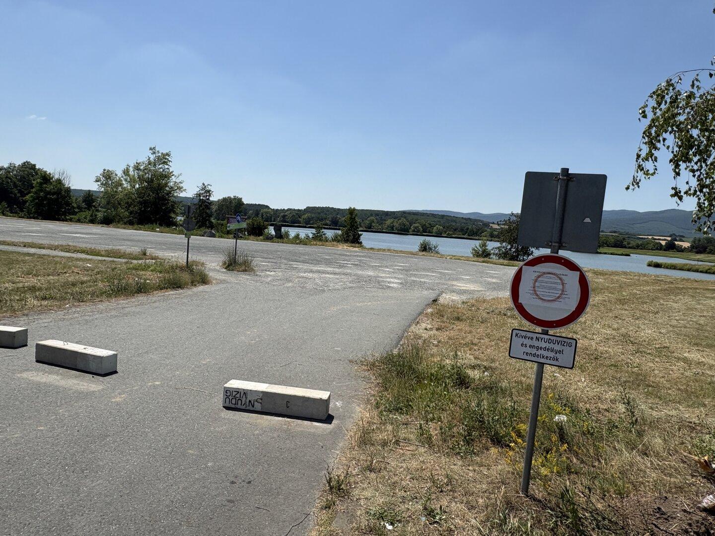 Sperrschilder blockieren eine Straße am Seeufer mit Blick auf Wasser und Hügel.