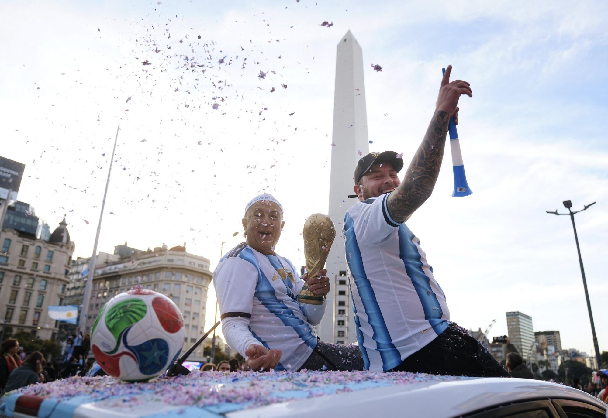Zwei Fans in Argentinien-Trikots feiern mit dem WM-Pokal vor dem Obelisken in Buenos Aires inmitten von Konfetti.