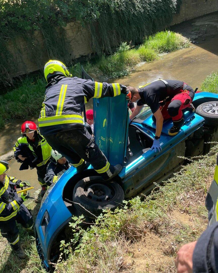 Feuerwehrkräfte retten eine Person aus einem auf der Seite liegenden blauen Auto am Bachufer.