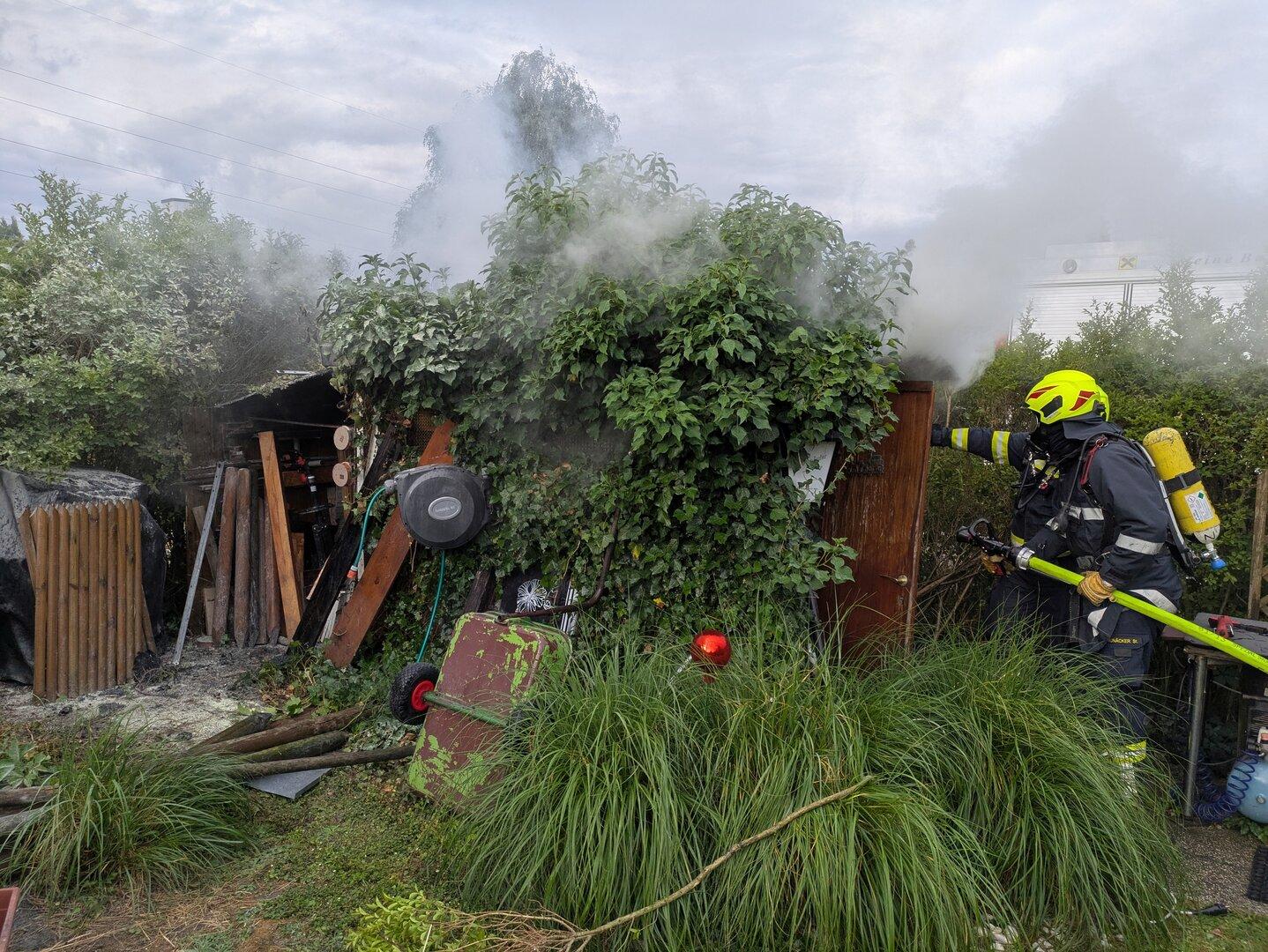 Feuerwehrmann löscht Rauch an einem stark bewachsenen Schuppen im Garten.