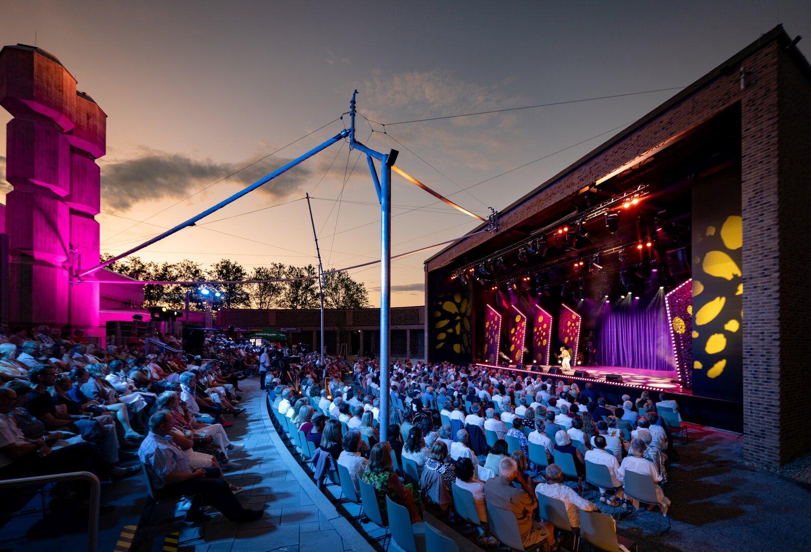 Open-Air-Theater bei Sonnenuntergang mit großem Publikum vor einer beleuchteten Bühne.