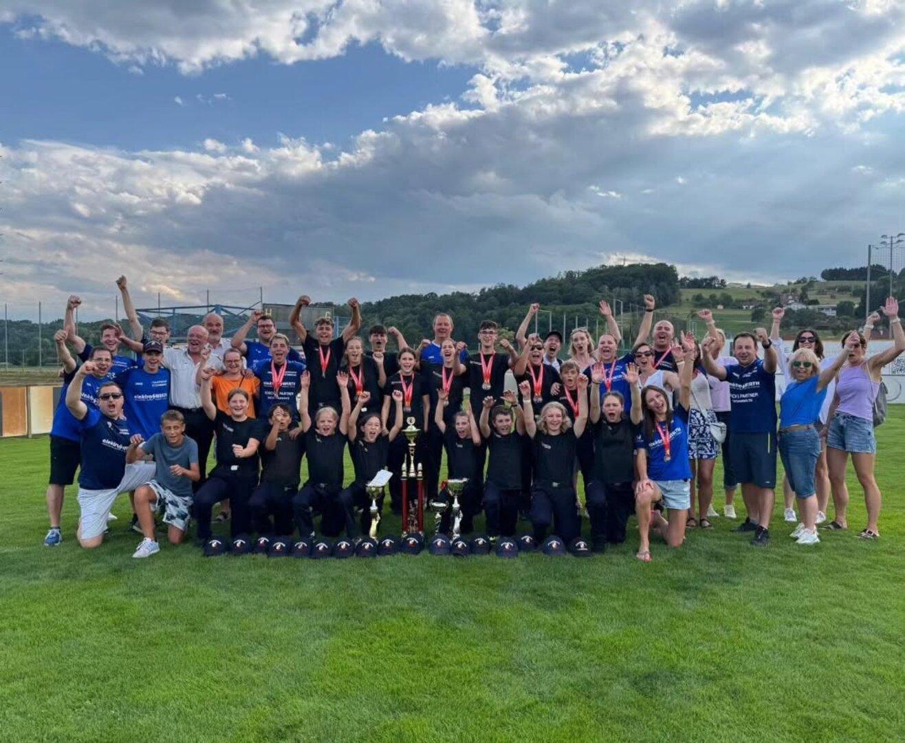 Große Gruppe jubelt auf einem Sportplatz mit Pokalen und Medaillen vor wolkigem Himmel.