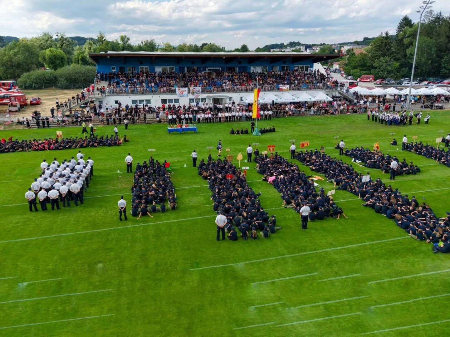 Zahlreiche Feuerwehrleute in Uniform sitzen in Gruppen auf einem Sportplatz vor einer voll besetzten Tribüne bei einer feierlichen Veranstaltung.
