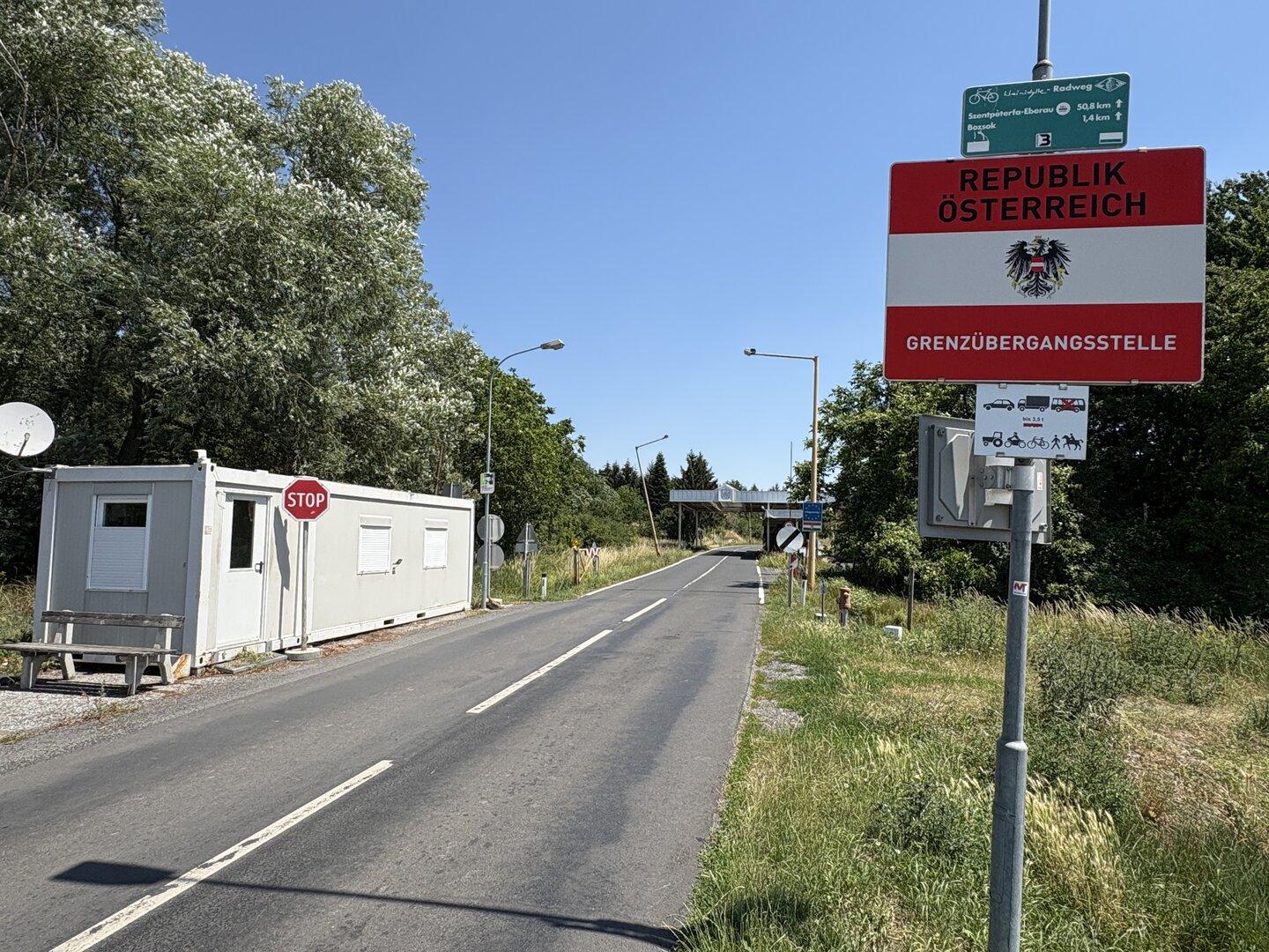 Grenzübergang nach Österreich mit Straßenschild, Kontrollhäuschen und leerer Landstraße unter klarem Himmel.