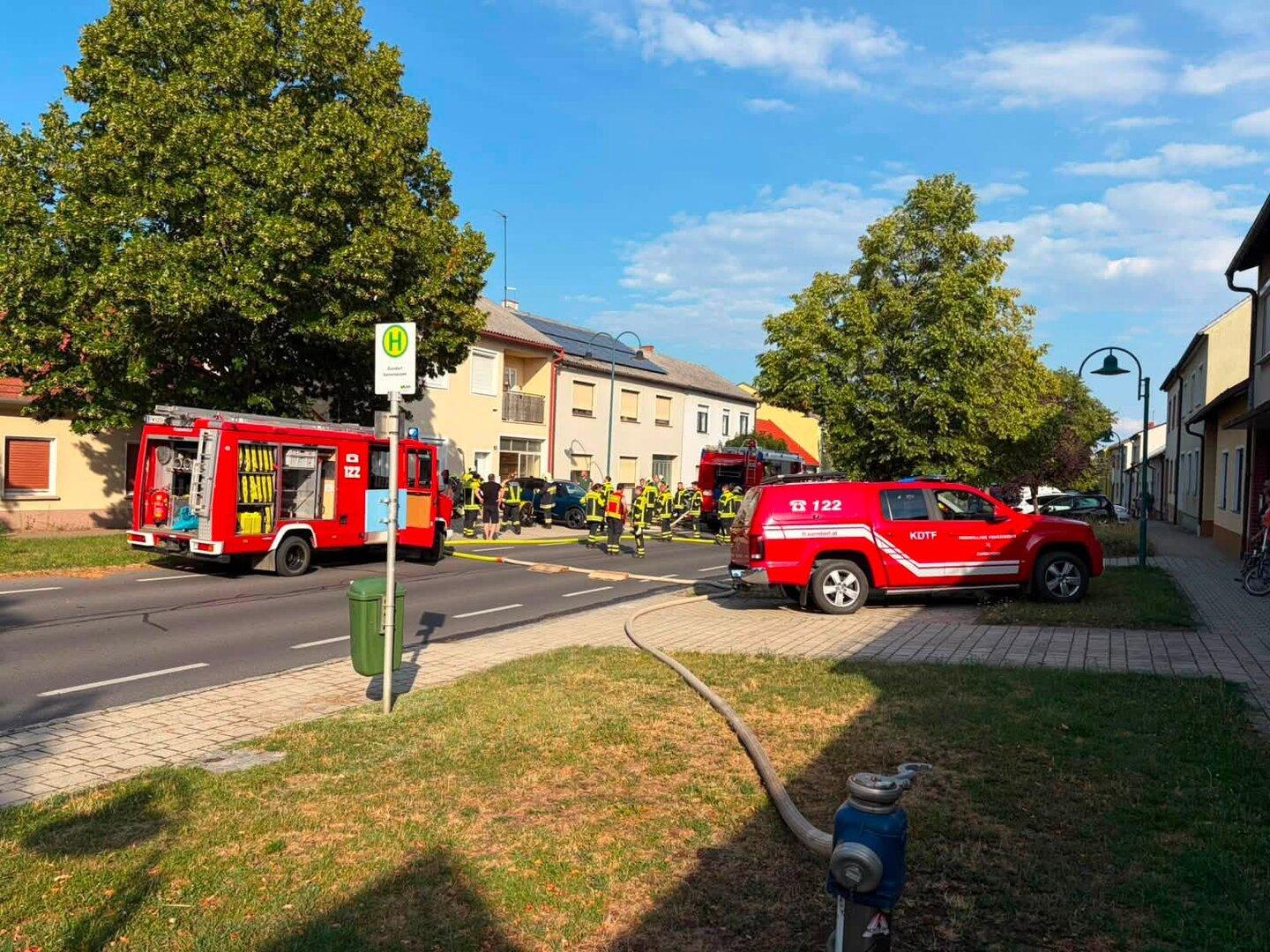 Feuerwehrkräfte und Einsatzfahrzeuge sichern eine Straße mit ausgelegtem Schlauch vor Wohnhäusern.