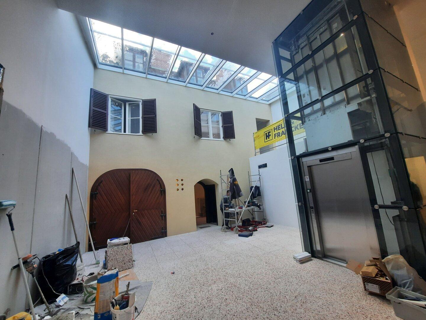 Innenhof mit Glasdach, alten Fenstern, großer Holztür und Aufzugsschacht während Renovierungsarbeiten.