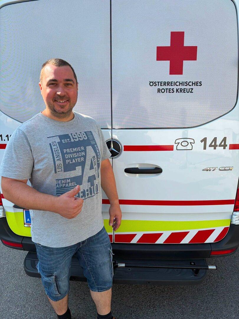 Mann lächelt vor einem Rettungswagen des Österreichischen Roten Kreuzes und zeigt den Daumen hoch.