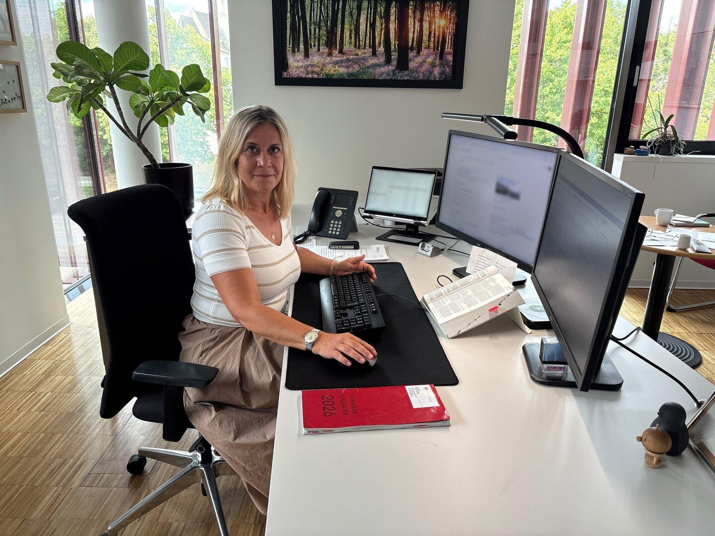 Frau sitzt an einem modernen Schreibtisch im Büro mit zwei Monitoren und Tastatur, umgeben von Pflanzen und Tageslicht.