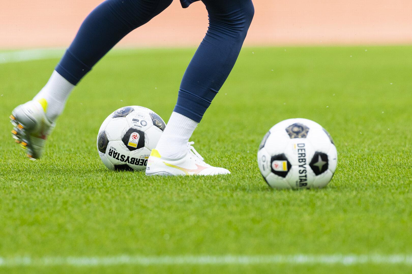 Fußballspiel mit zwei Bällen und den Beinen einer Spielerin auf dem Rasen.