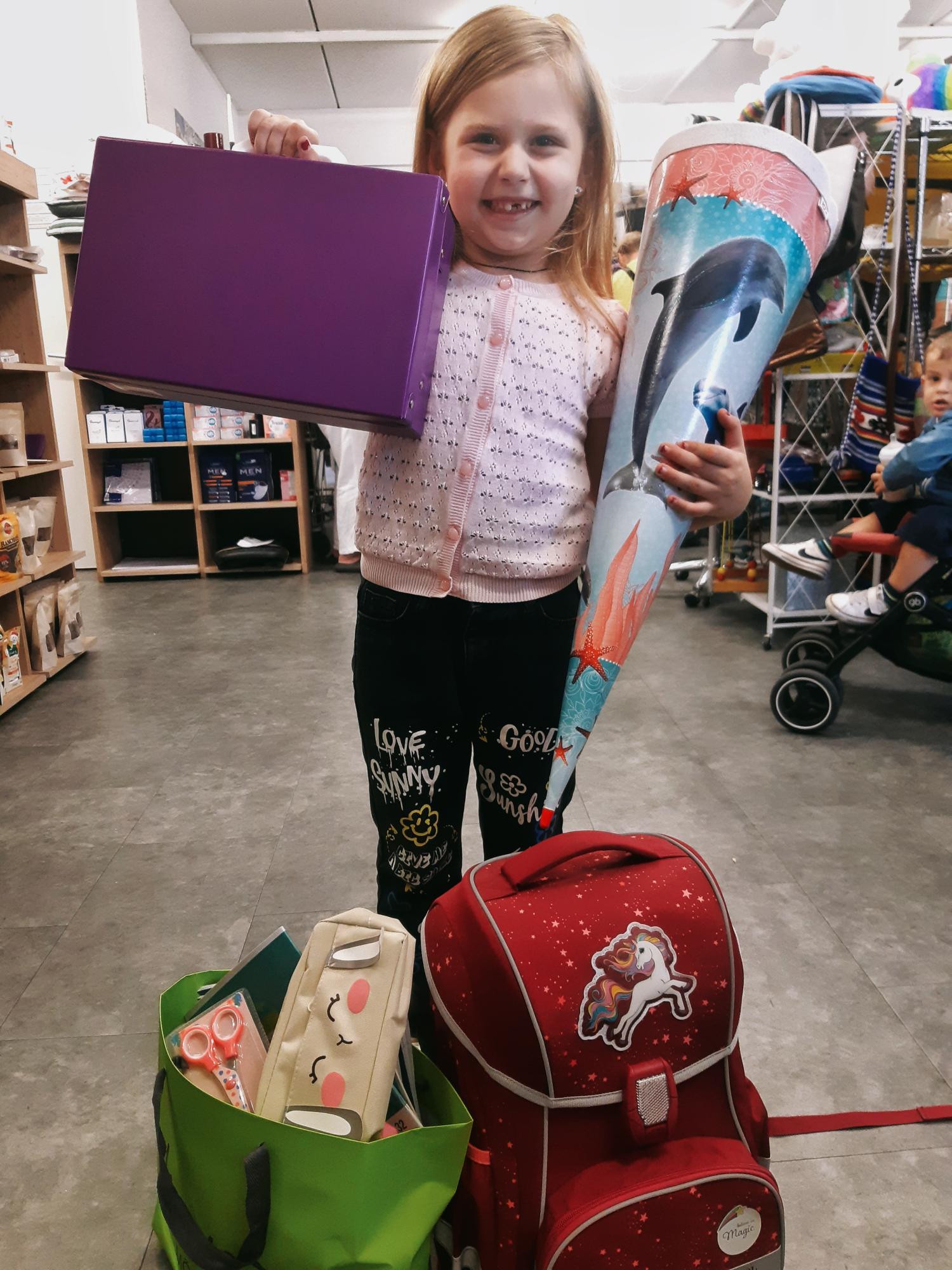 Mädchen lächelt und hält einen lila Ordner sowie eine Delfin-Schultüte in einem Laden.