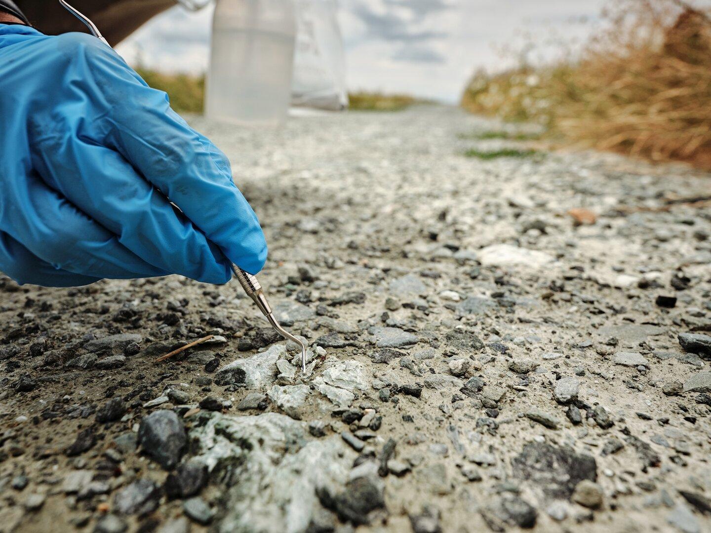 Behandschuhte Hand untersucht mit einem Metallwerkzeug den rissigen Asphalt am Straßenrand.