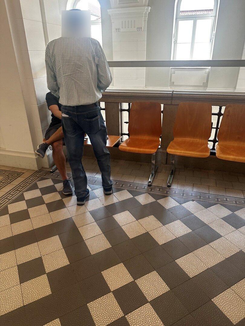 Zwei Männer stehen in einem hellen Flur neben einer Sitzreihe mit Holzstühlen.