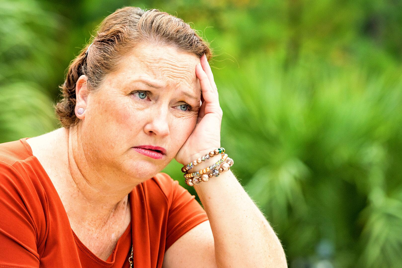 Eine nachdenklich wirkende Frau mit rötlichem Haar und orangefarbenem Oberteil stützt ihren Kopf nachdenklich gegen die Hand.