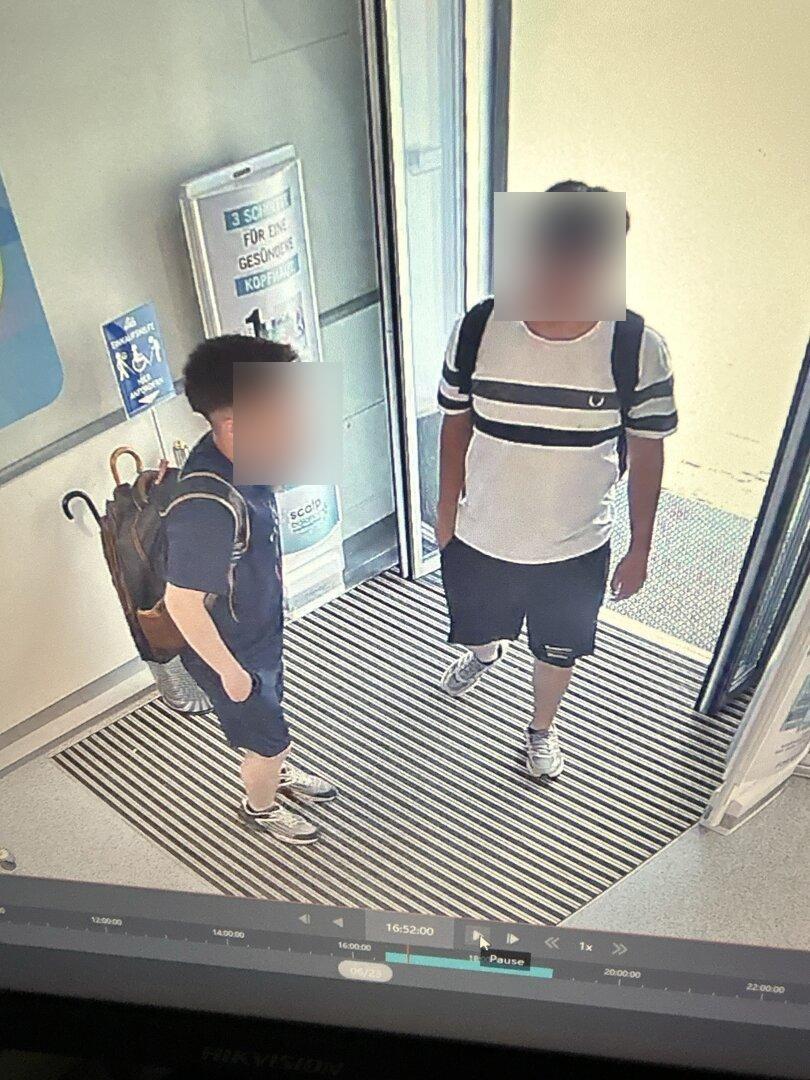 Zwei junge Männer mit Rucksäcken stehen im Eingangsbereich vor einer Glastür.