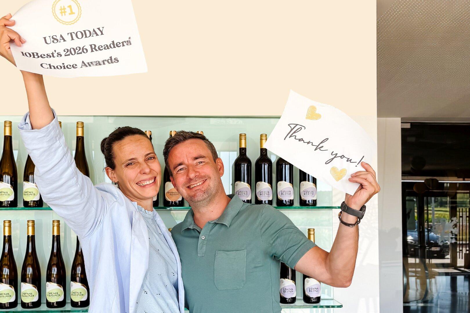 Ein lächelndes Paar hält Schilder mit der Aufschrift „USA TODAY 10Best's 2026 Readers' Choice Awards“ und „Thank you!“ vor einem Weinregal.
