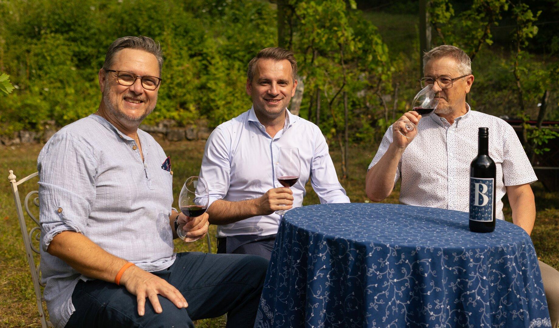 Drei Männer sitzen draußen an einem Tisch, trinken Rotwein und lächeln in die Kamera.