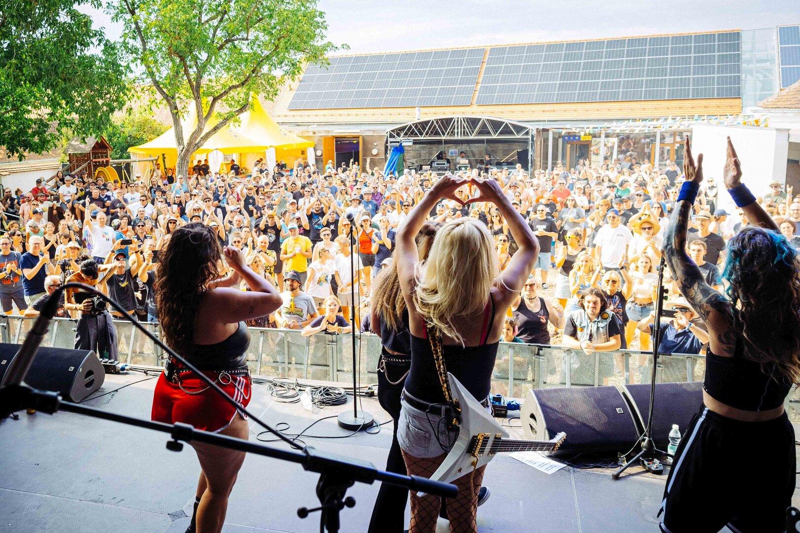 Band auf einer Open-Air-Bühne vor jubelnder Menschenmenge, die Sängerinnen begrüßen das Publikum.