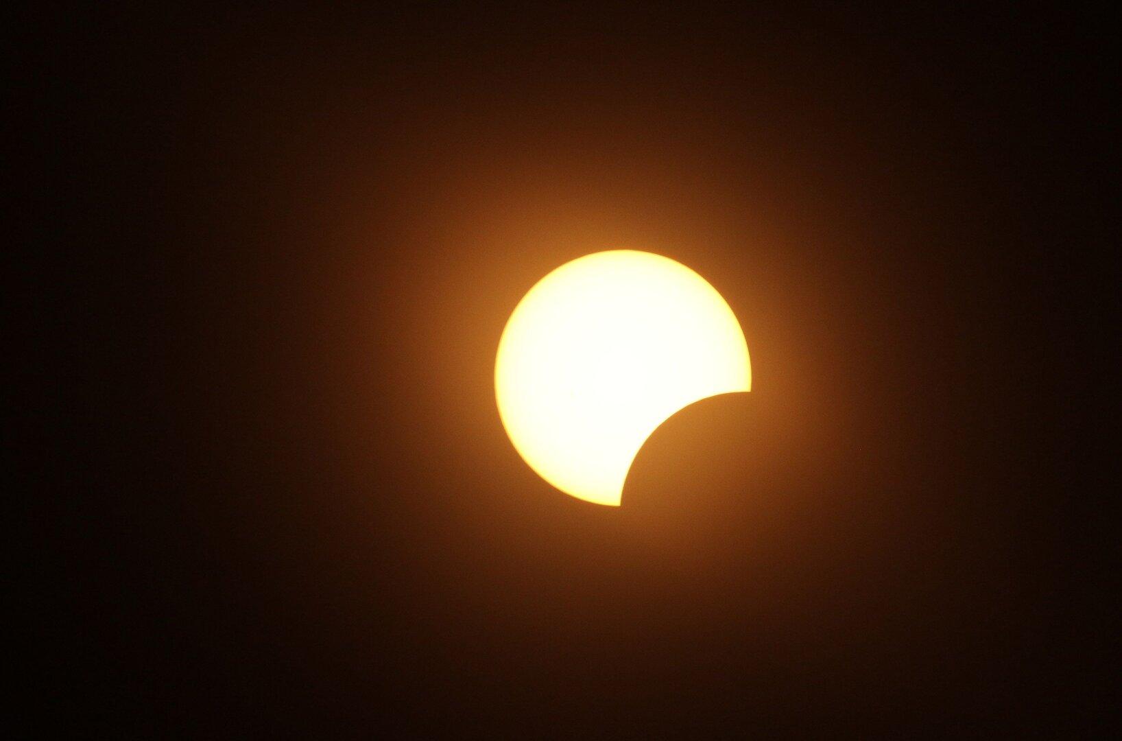 Teilweise verfinsterte Sonne mit dunkler Sichel vor orangefarbenem Himmel.