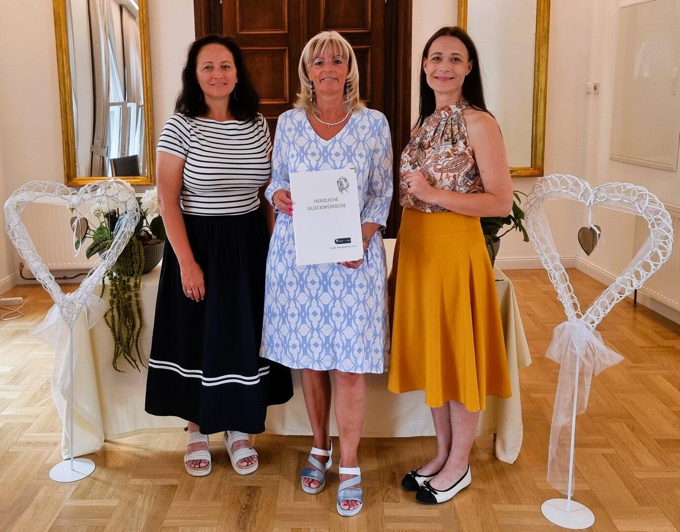 Drei Frauen (in der Mitte Stadtchefin Böhm) stehen nebeneinander, wobei die mittlere eine Urkunde der Stadt Neusiedl am See hält, flankiert von zwei weißen Herz-Dekorationen.